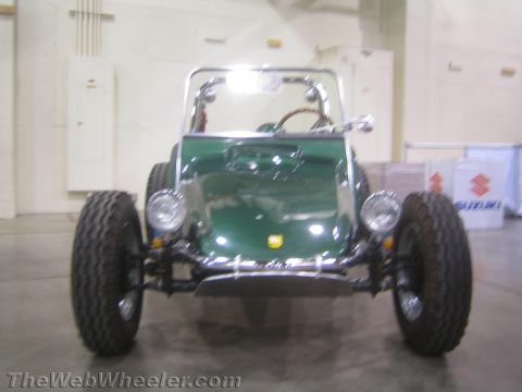 1990 Ford Bronco - green fiberglass dune buggy with chrome roll bar and round headlights sits facing the camera on an indoor show floor near a Suzuki banner.