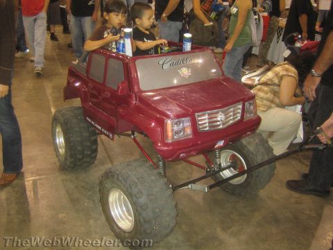1990 Ford Bronco - Two small children sit in a maroon Cadillac-badged kids' ride-on car built onto oversized off-road wheels at an indoor event.