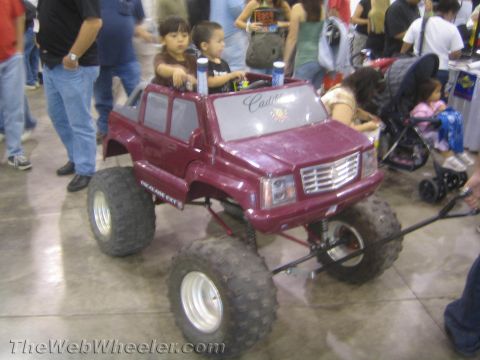 1990 Ford Bronco - maroon Cadillac Escalade-style children's ride-on toy raised on big off-road tires, with kids aboard at an indoor event.