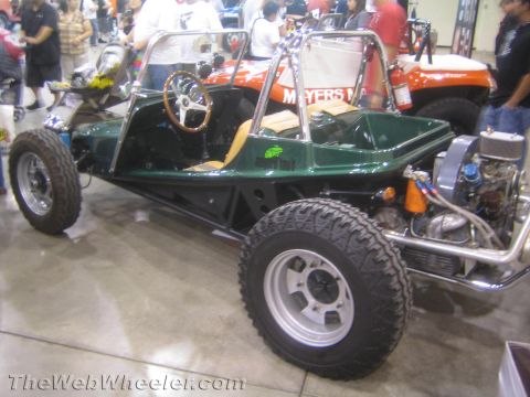 1990 Ford Bronco - dark green dune buggy with tan upholstery, a wood-rim steering wheel and a rear-mounted engine sits on display at an indoor show.