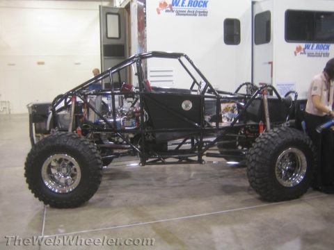 1990 Ford Bronco - black tube-frame rock crawler buggy on chrome beadlock wheels and large tires displayed indoors next to a W.E. Rock trailer.