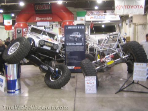 1990 Ford Bronco - Two tube-frame rock crawlers posed on display stands beneath a red Tecate arch, with an info placard and trophies at an indoor event.
