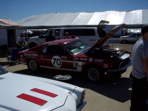 1971 Ford Mustang - A red 1969 race car with sponsor decals and the number 70 is parked next to a white car under tents.