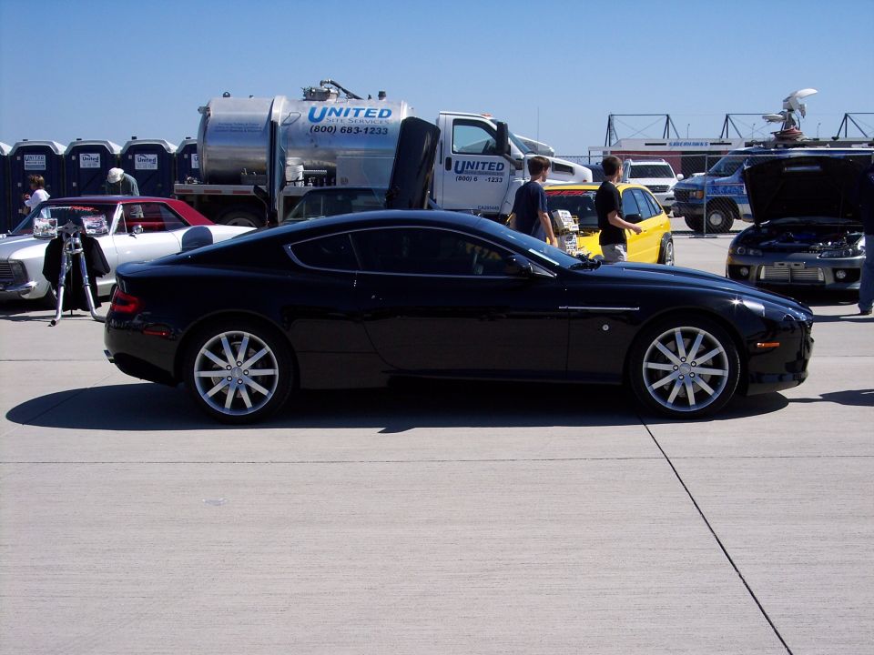Black coupe with silver multi-spoke wheels parked on concrete, other vehicles and portable toilets in background
