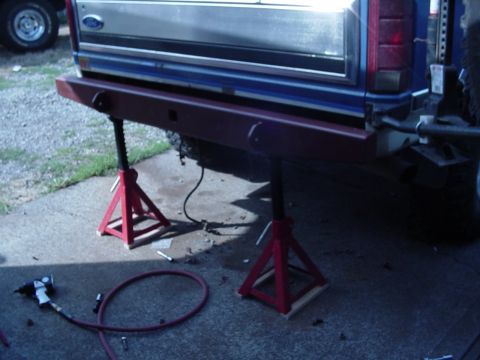 1985 Ford Bronco - A red aftermarket bumper is installed on a blue Ford truck, which is raised on jack stands.