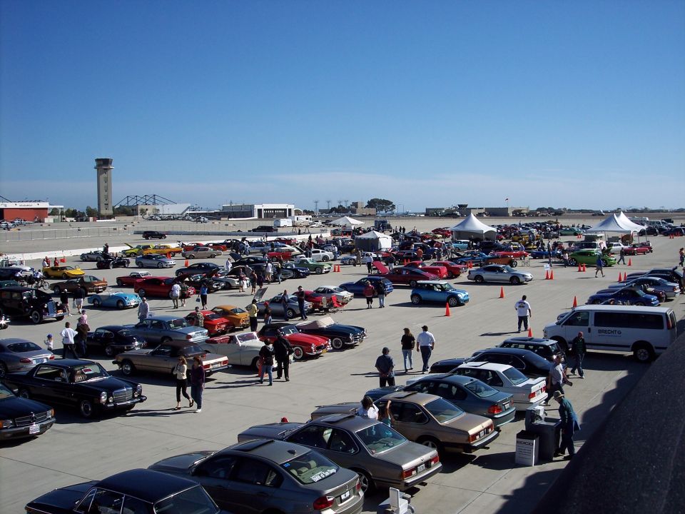 Outdoor car show with dozens of cars parked on asphalt under a clear blue sky. People are milling around the vehicles