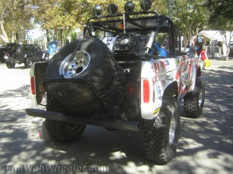 1990 Ford Bronco - Rear three-quarter view of a white off-road race truck carrying a spare tire, sticker-covered tailgate and a roof-mounted light bar.