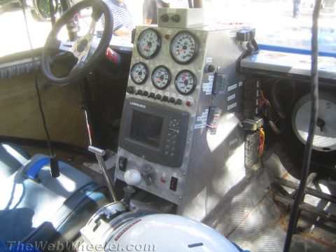 1990 Ford Bronco - Bare-metal race-truck interior with a steering wheel, stacked gauges, a Lowrance navigation unit and a helmet resting on the seat.