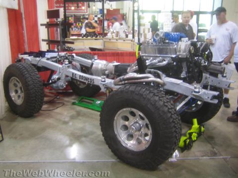 1990 Ford Bronco - BC Broncos rolling chassis fitted with a chrome-dressed V8 engine, axles and large off-road tires displayed on an indoor show floor.