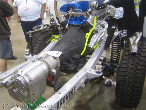 1990 Ford Bronco - Detail of a bare frame with a blue-intake V8 engine, transmission, coilover shocks and an off-road tire at an indoor display.