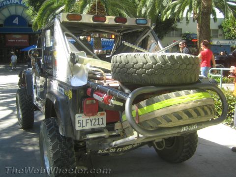 1990 Ford Bronco - Rear three-quarter view of a lifted Jeep with a tube bumper, spare tire, winch and California plate 5LFY211 among palm trees at an event.