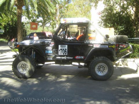 1990 Ford Bronco - Side profile of a black lifted Jeep race rig numbered 1706 on large off-road tires with side exhaust, parked outdoors near palms.