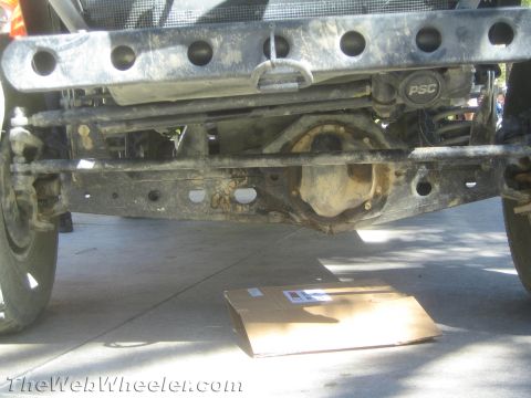 1990 Ford Bronco - Low front view beneath an off-road rig showing the front axle, drilled crossmember and a PSC-labeled steering component over a parts box.
