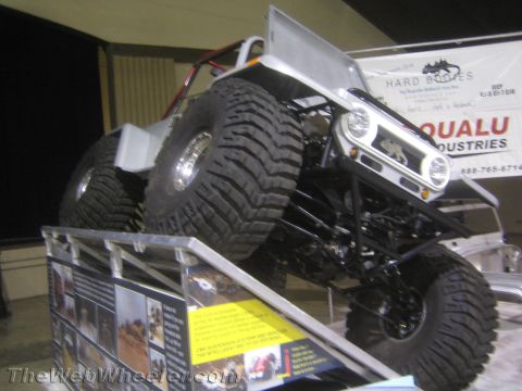 1990 Ford Bronco - silver-bodied off-road buggy on huge knobby tires climbing a metal ramp on a display stand under a Hard Bodies / Qualu Industries banner.