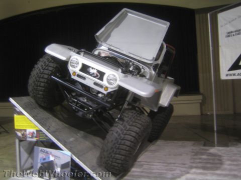 1990 Ford Bronco - silver unpainted rock-crawler buggy with round headlights and chunky off-road tires displayed on an angled ramp at an indoor expo.