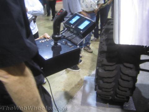 1990 Ford Bronco - black handheld control box with a joystick, switches and two small LCD screens, held beside a large off-road tire at an indoor expo.