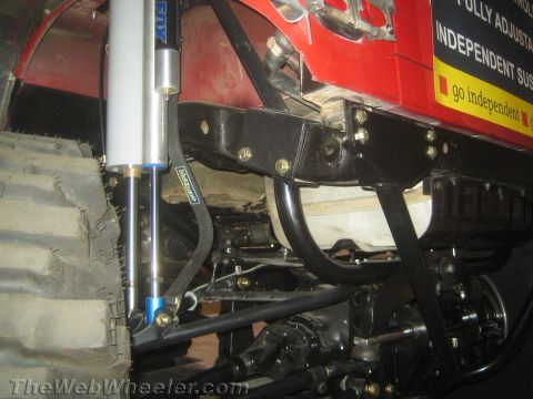 1990 Ford Bronco - Underside of a red lifted vehicle showing a FOX coilover shock, suspension control arms and frame against an off-road tire.