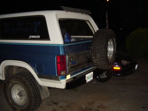 1985 Ford Bronco - Rear view of a blue and white Ford Bronco with a spare tire, parked next to a dark sedan at night.