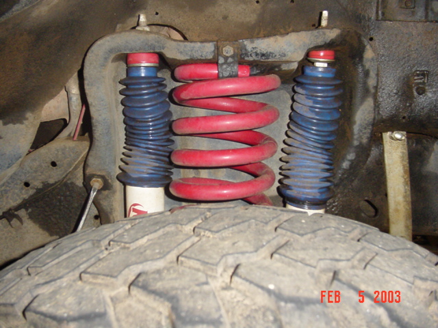 Red coil spring and dual shocks with blue boots in front suspension above a tire