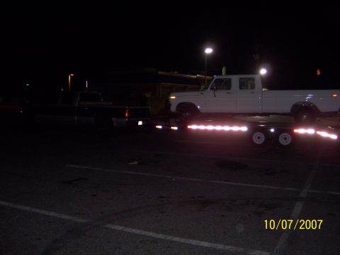 1978 Ford Bronco - White crew cab pickup truck being transported on a trailer at night, with another truck in the background.