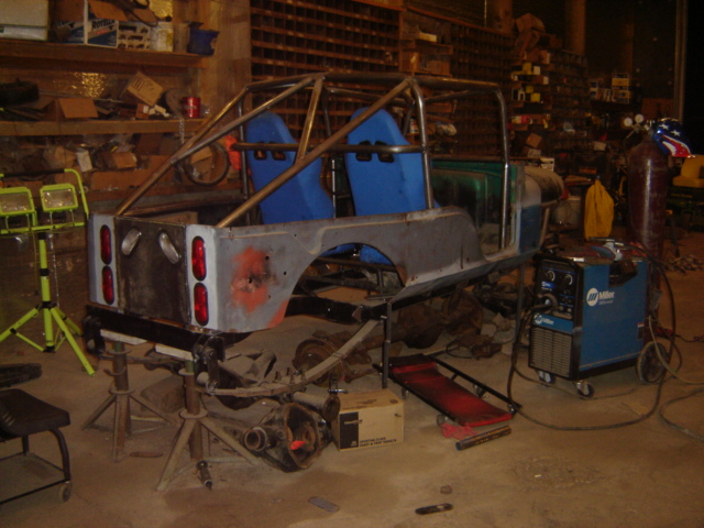 Custom vehicle chassis with roll cage and blue seats, in a workshop with welding equipment