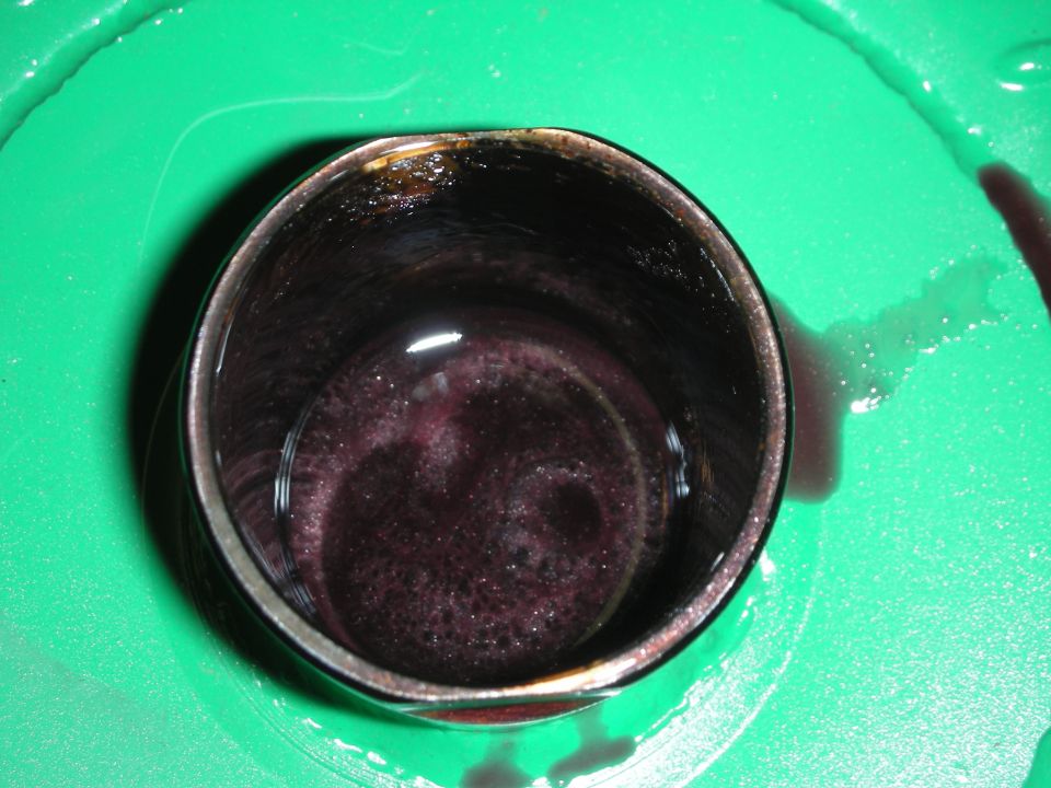 Close-up of dark purple fluid with bubbles in a metal cylinder, sitting on a green surface with some spilled fluid