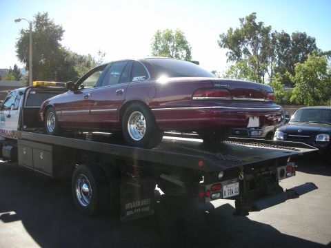 1992 Ford Crown Victoria - A 1992 Ford Crown Victoria is loaded onto a flatbed tow truck, with a dark SUV parked behind.