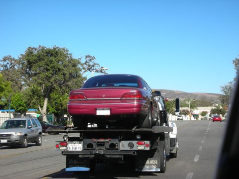 1992 Ford Crown Victoria - A red sedan is loaded onto a tow truck, heading down a sunny street with other vehicles visible.