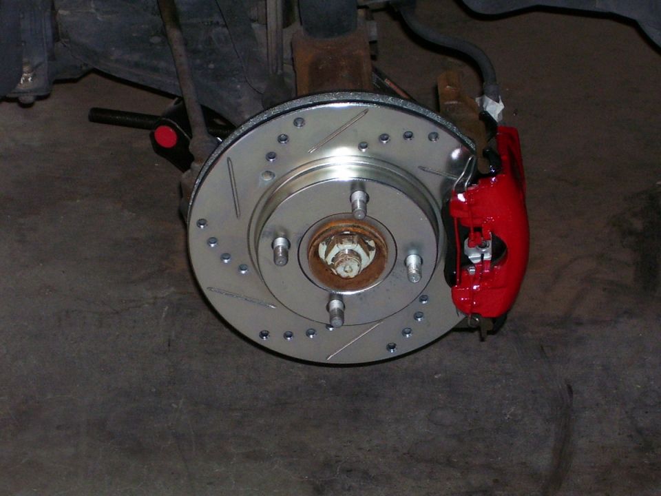 Drilled and slotted brake rotor with red caliper installed on hub, wheel removed, in a garage
