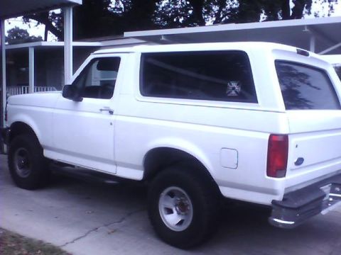 1989 Ford Bronco - Three-quarter rear view of a white full-size two-door Ford Bronco with removable hardtop, parked on a driveway beside a carport. It wears f…