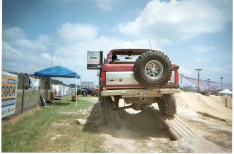 1995 Ford Bronco - Rear view of a lifted red and white Ford Bronco climbing a dirt obstacle course mound at an outdoor event, with a swing-out spare tire carr…