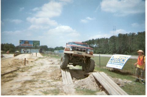 1995 Ford Bronco - A heavily modified pickup truck navigates an off-road obstacle course under a partly cloudy sky.