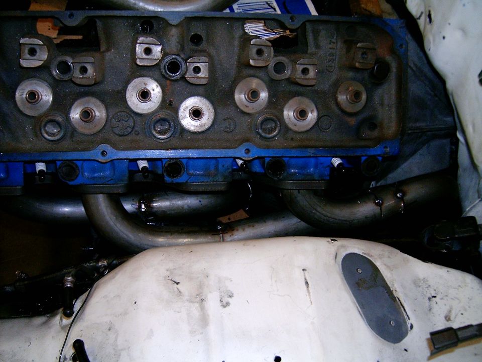 Engine cylinder head with blue valve covers and attached exhaust headers. Spark plugs are visible