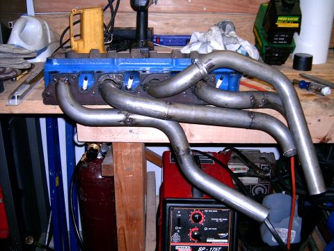1978 Ford Mustang II - Exhaust headers are shown attached to a blue engine block, with various tools and equipment visible on the workbench.
