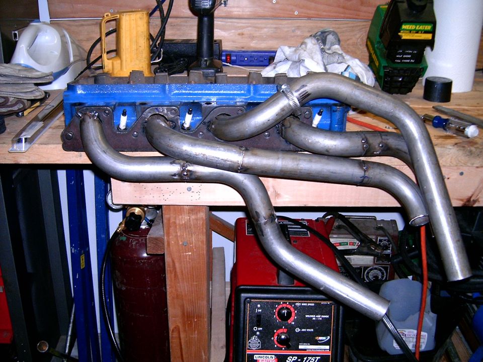 Exhaust headers attached to a blue engine block on a workbench, with welding equipment in the background