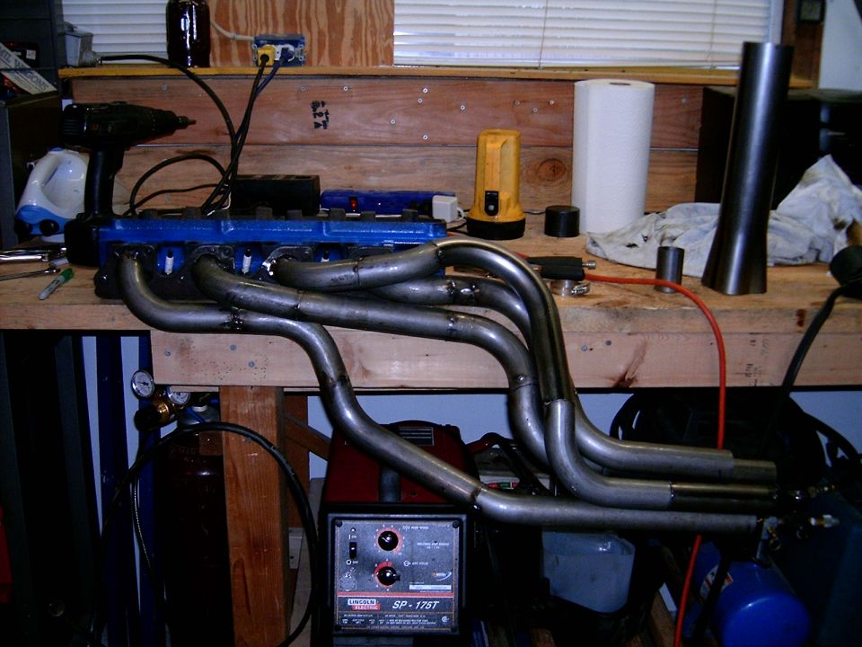 Headers on a workbench with a blue engine block and welding equipment
