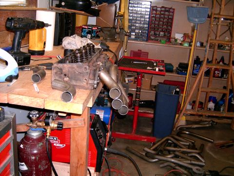 1978 Ford Mustang II - An engine cylinder head with exhaust headers is on a workbench, with tools and welding equipment nearby.