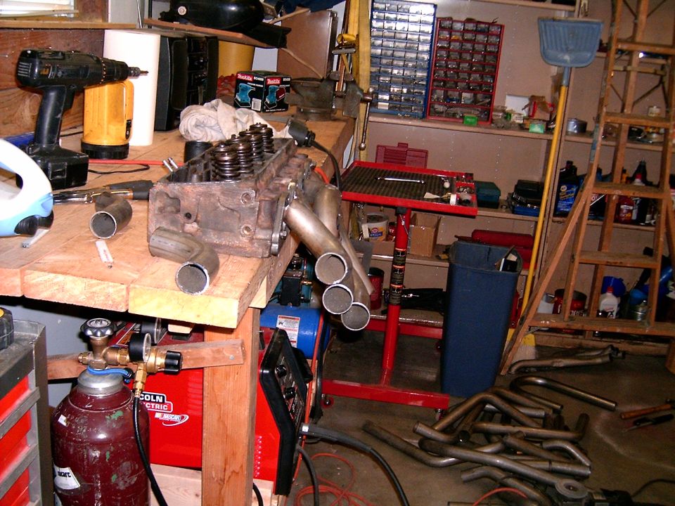 Engine cylinder head with exhaust headers on a workbench next to a welding machine