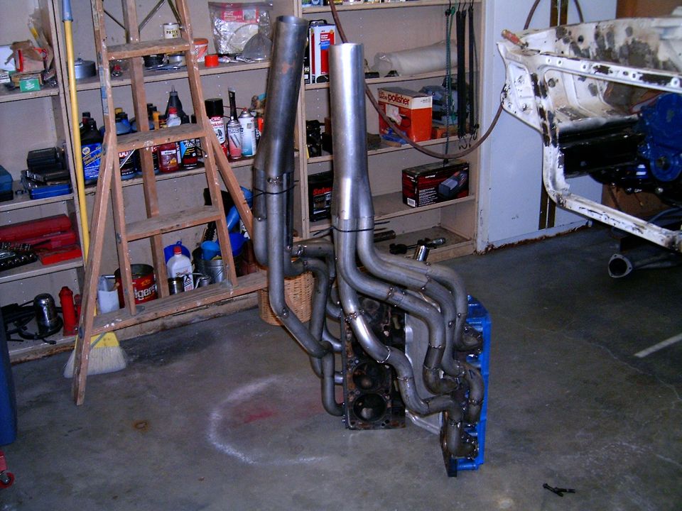 V8 engine block with custom exhaust headers installed, sitting on a blue pallet in a garage