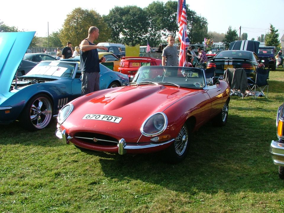 Red convertible sports car with license plate 670 PDT parked on grass. Another blue car with hood up is to the left