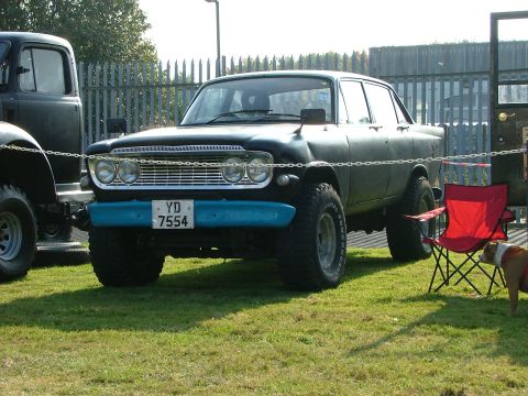 1988 Ford Sport - This modified sedan features large off-road tires and a bright blue bumper, parked on grass next to another vehicle.
