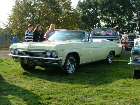 1988 Ford Sport - A pale yellow 1965 Chevrolet Impala convertible is displayed on a grassy field, with other vehicles visible in the background.