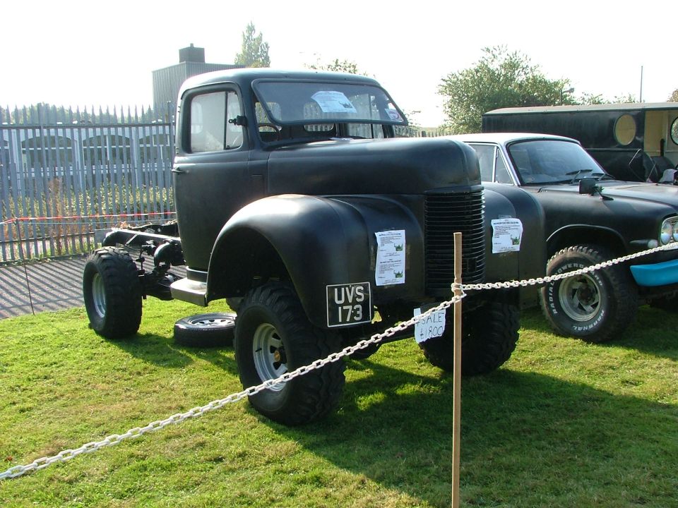 Black truck chassis with large tires, parked on grass next to another car