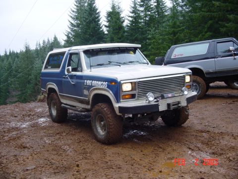 1985 Ford Bronco - Two Ford Broncos are parked on a muddy trail surrounded by trees.