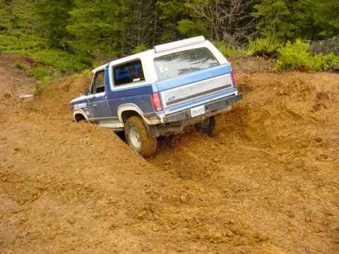 1985 Ford Bronco - A blue and white Ford Bronco tackles a challenging muddy terrain during an off-road event.
