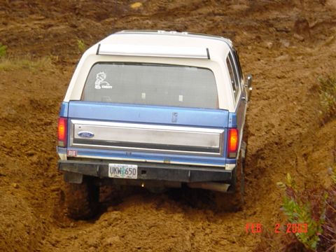 1985 Ford Bronco - Ford Bronco navigates a muddy off-road course, showing its rear end and license plate.