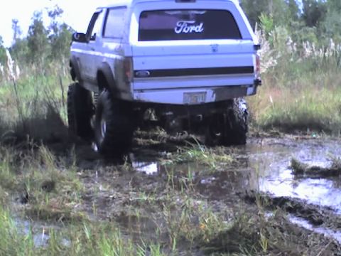 1995 Ford Bronco - A lifted 1995 Ford Bronco navigates a muddy off-road trail, kicking up water and mud.