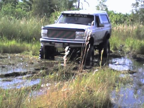 1995 Ford Bronco - This lifted SUV is navigating through a wet, grassy terrain with visible mud and water.