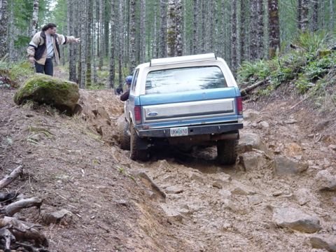 1985 Ford Bronco - A man points towards a blue and white Ford Bronco as it drives through a challenging off-road terrain.