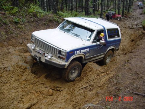 1985 Ford Bronco - A 1985 Ford Bronco tackles a challenging muddy obstacle during an off-road event, with another vehicle visible in the background.